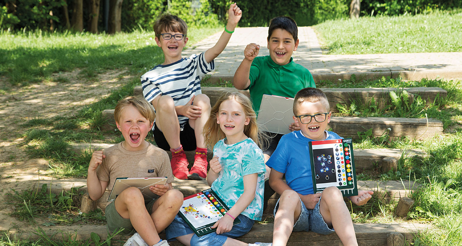 Lernen mit LOGICO macht Spaß Jubelnde Kinder sitzen draußen auf einer Treppe auf dem Schulgelände und lernen mit LOGICO