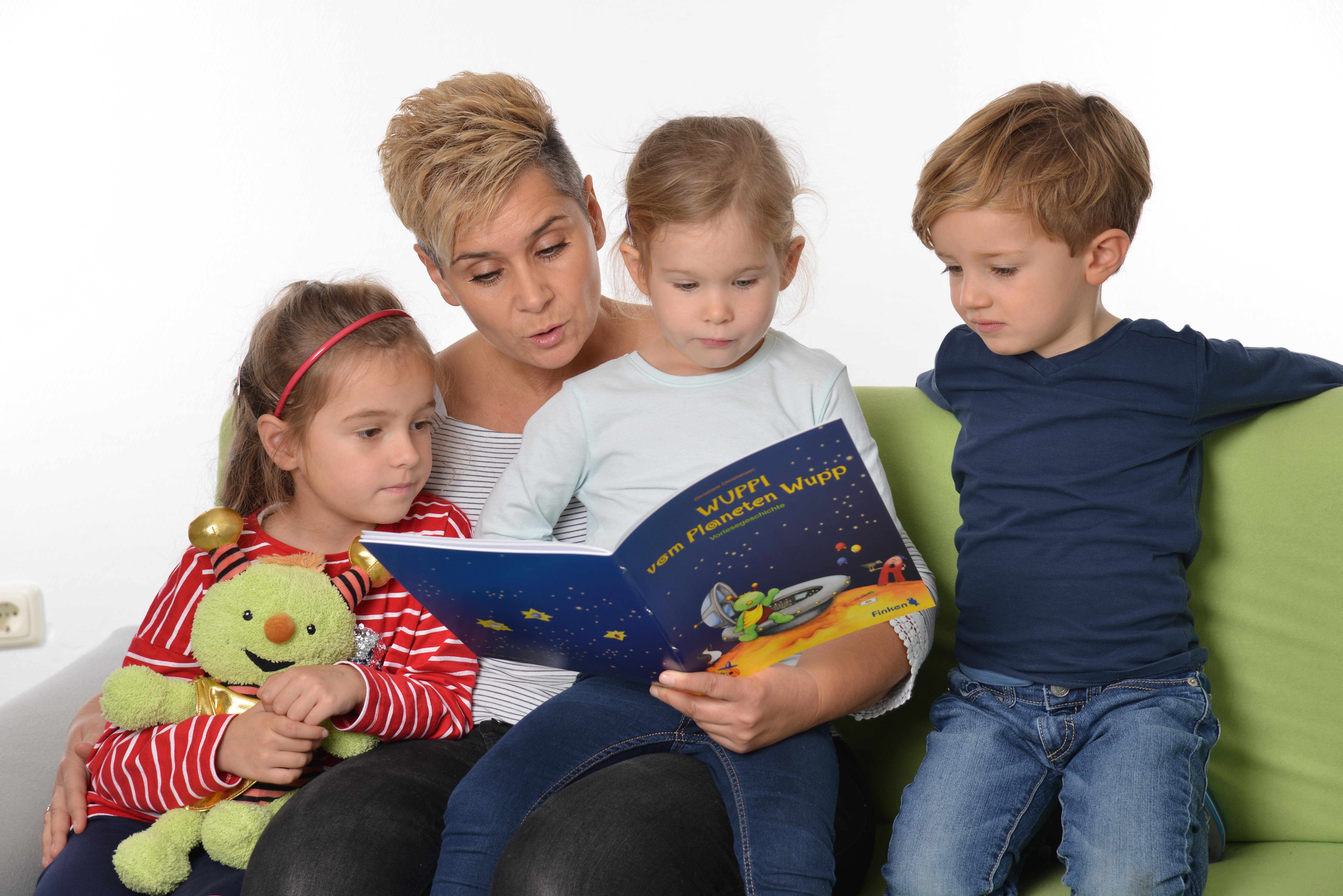 Erzieherin sitzt mit Kindern auf einem grünen Sofa und liest eine Geschichte von Wuppi vor. Ein Kind hält die Handpuppe "Wuppi" im Arm